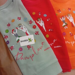 Varias sudaderas de algodón orgánico con estampación de dantzaris en colores rojo, verde y naranja, diseño exclusivo de ButtiBou Pamplona. Multiple organic cotton sweatshirts with Dantzaris print in red, green, and orange, exclusive design by ButtiBou Pamplona.